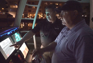 A man in a black polo pointing at a navigation screen on a boat while a man in a blue polo looks ahead and drives