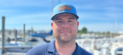 Robert Finch wearing a blue cap with text and a navy polo shirt standing by a sunny marina.